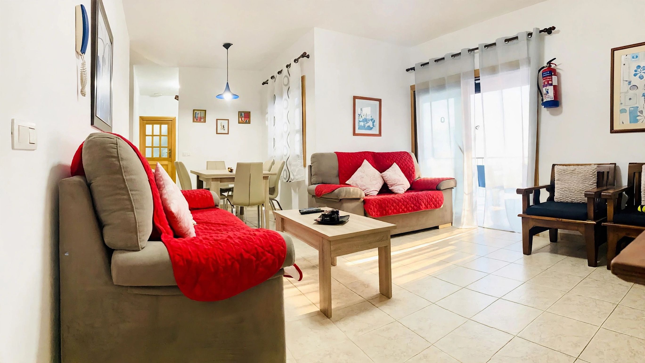 Een woonkamer met een bank, een bijzettafeltje en een rode plaid. Er is een eethoek met een tafel en stoelen. In de kamer hangt een brandblusser aan de muur.