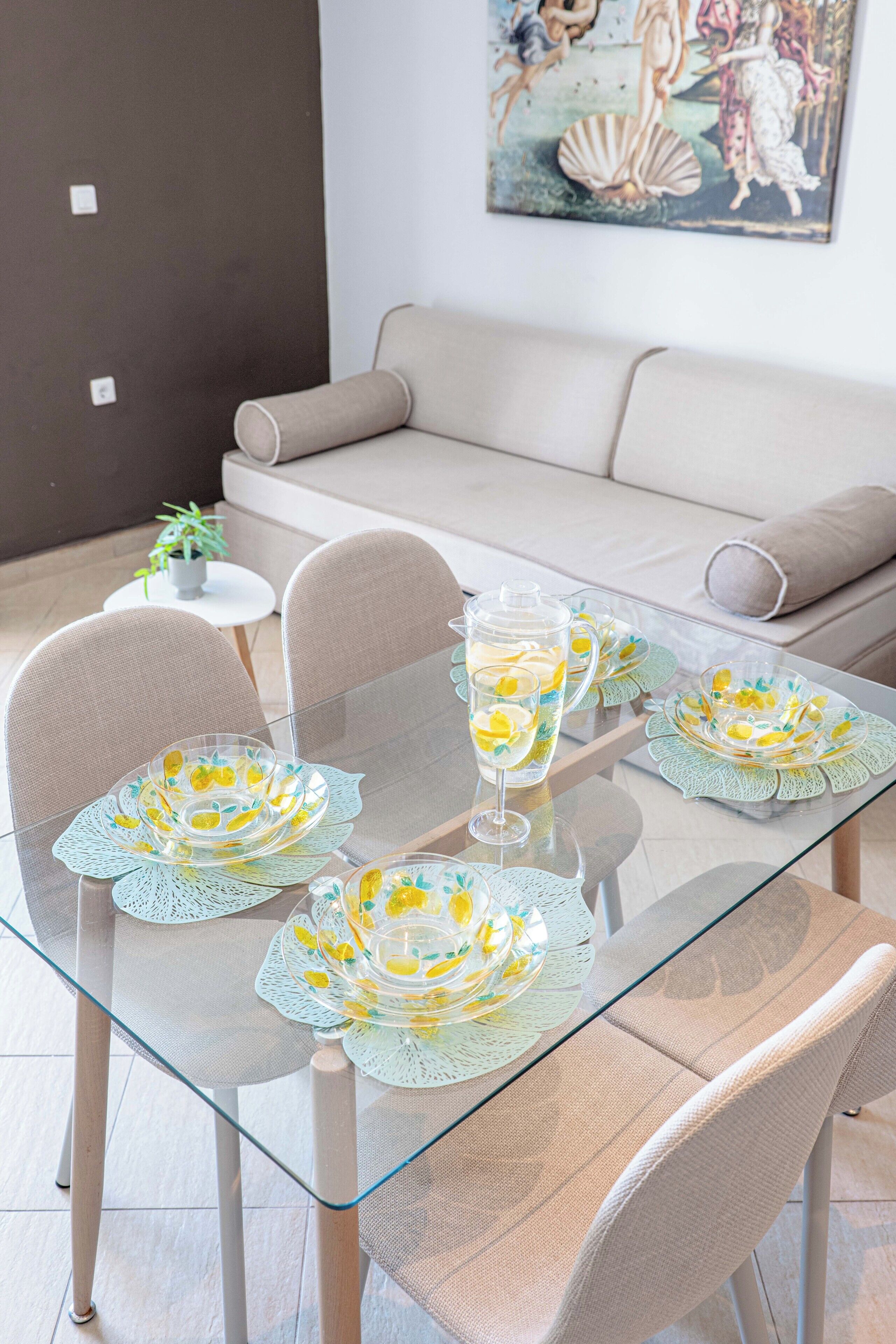 Ett matbord med glasskiva, dekorerat med blommig bordsdekoration, en kanna och koppar, placerat i ett modernt vardagsrum med en soffa och ett litet runt bord med en växt.