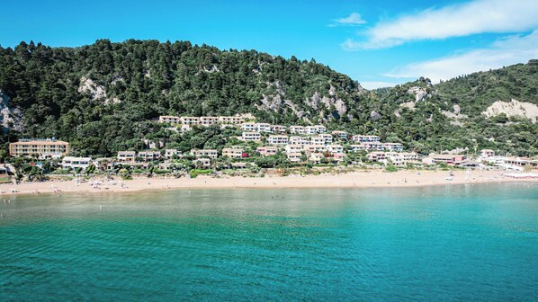 Un pueblo costero con hoteles frente a la playa, aguas turquesas cristalinas y una colina cubierta de vegetación densa.