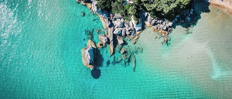 Vue aérienne d’une zone côtière avec des eaux turquoise limpides, des affleurements rocheux et une plage où se trouvent des personnes.