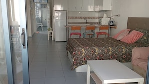 Une chambre compacte comprenant un lit, une table de chevet et un petit bureau avec une chaise. On aperçoit, au fond, une kitchenette équipée de placards et d’un réfrigérateur.