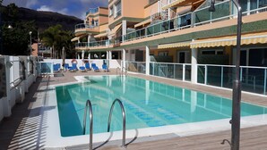 Une piscine à l’eau d’un bleu cristallin, entourée d’une terrasse en bois et jouxtant un immeuble à plusieurs étages doté de balcons.