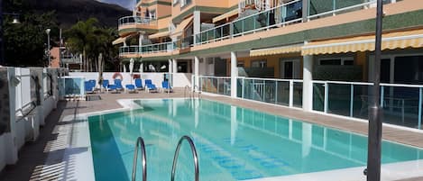 A swimming pool with a clear blue water, surrounded by a wooden deck, and adjacent to a multi-story building with balconies.