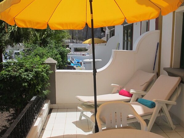 Eine sonnendurchflutete Terrasse mit zwei weißen Liegestühlen, einer mit einem rosa Kissen und der andere mit einem blauen Handtuch. Ein gelber Sonnenschirm spendet Schatten.