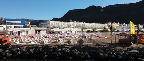 A beachside hotel with rows of sun loungers and umbrellas, a rocky shoreline, and buildings in the background.