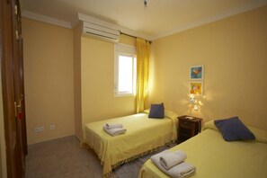 A room with two beds, a window with curtains, a wall-mounted air conditioner, and a nightstand with a lamp.