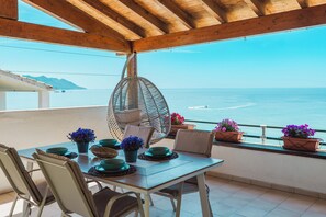 Une terrasse dotée d’un plafond en bois, une table décorée de bols verts et de plantes en pot, et une vue sur la mer.