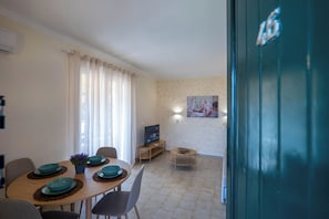 Een eetgedeelte met een tafel voor vier personen, een televisie op een standaard en een schilderij aan de muur.