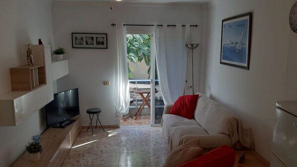 A living room with a TV, a sofa, a small table, and a balcony with a view of greenery.