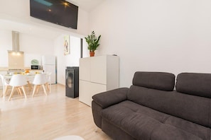 A modern living room with a grey sofa, a flat-screen TV mounted on the wall, a dining area with white chairs and a table, and a kitchen area with a refrigerator and microwave.