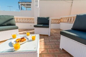 A rooftop terrace with wicker furniture, a table with food and drinks, and a clear view of the sky.