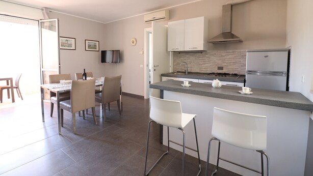 Una cocina moderna con una isla central, armarios blancos y electrodomésticos de acero inoxidable. Cuenta con un comedor con sillas y una mesa.