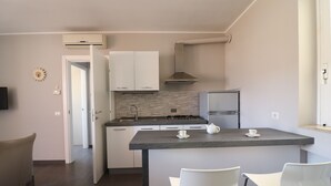Una cocina moderna con gabinetes blancos, estufa a gas y refrigerador de acero inoxidable. Hay una pequeña isla con dos sillas blancas y una tetera encima.