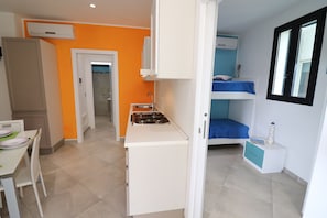 Une cuisine compacte équipée d’une cuisinière à gaz, d’une table à manger avec des chaises, d’un lit superposé avec un matelas bleu et d’une armoire blanche.