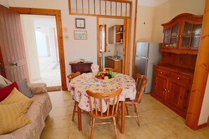 Un coin repas avec une table ronde, des chaises, un buffet en bois et une cuisine visible à travers une porte ouverte.