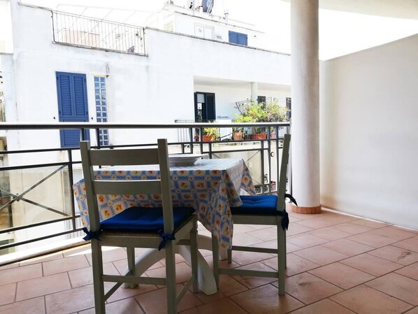 A balcony with a table and two chairs, a white building with blue shutters in the background, and potted plants.