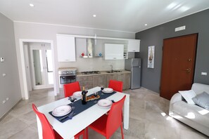 Une cuisine moderne avec des meubles blancs, une table à manger entourée de chaises rouges et un canapé gris dans l’espace salon.
