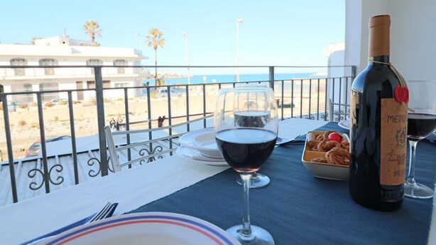 A table with a bottle of wine, a glass of red wine, and a bowl of cookies on a balcony overlooking a beach. - Casa Vanessa – Modern one-bedroom apartment with sea view in central Otranto (Otranto)