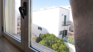 Ein Fenster mit Türgriff, Blick auf ein weißes Gebäude mit Balkon und ein grüner Busch im Vordergrund.