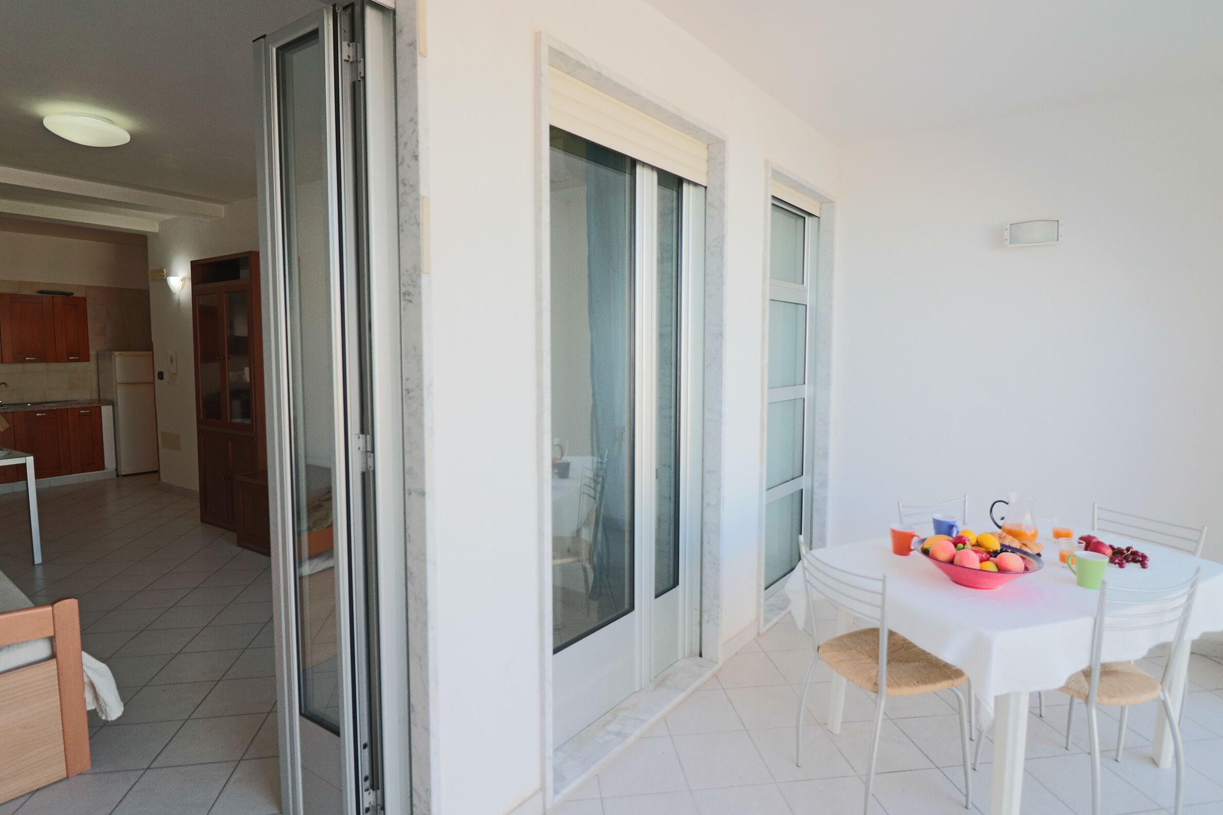 A dining area with a white table and chairs, a bowl of fruit, and a kitchen in the background.