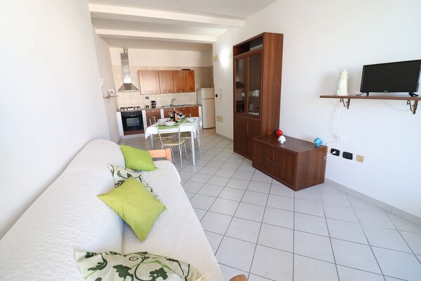 A modern living room with a sofa, a dining area with a table and chairs, a kitchen with cabinets and appliances, and a TV on a shelf.