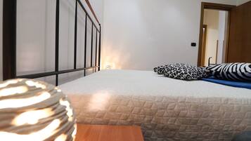 Quarto com cama, mesa de cabeceira de madeira, travesseiro estampado e porta que dá para outro cômodo.