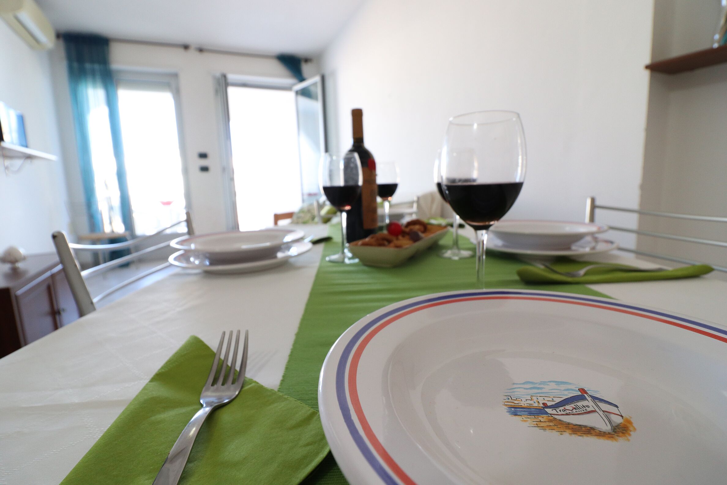 Uma mesa de jantar posta com pratos, copos e talheres, com uma garrafa de vinho e uma tigela de frutas ao fundo.