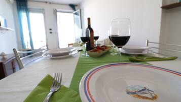 Uma mesa de jantar posta com pratos, copos e talheres, com uma garrafa de vinho e uma tigela de frutas ao fundo.