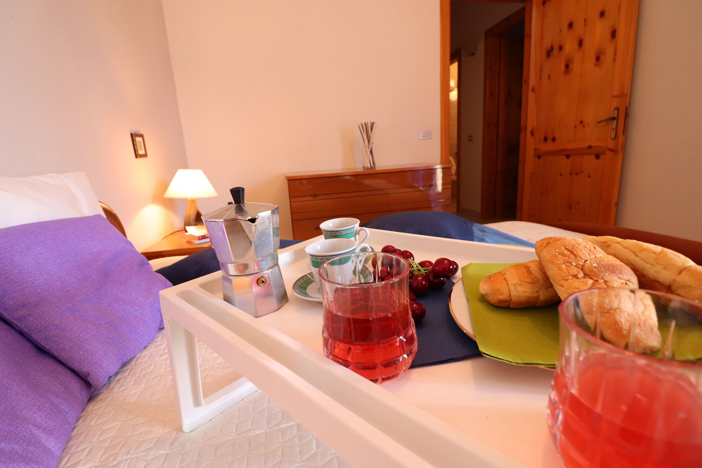 Un lit bien fait, avec un oreiller violet, une table de chevet avec une lampe, un plateau avec de la nourriture et des boissons, et une porte en bois en arrière-plan.