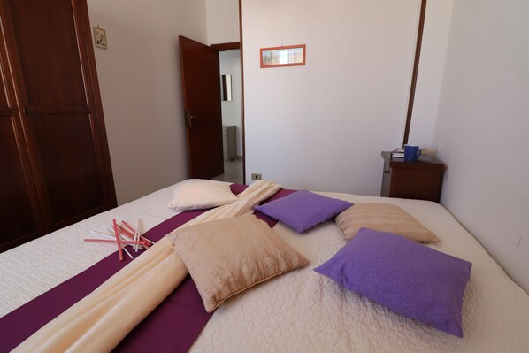 Un lit bien fait, recouvert d’une couvre-lit aux tons violet et beige, une table de chevet en bois avec une tasse bleue, et une armoire en bois.
