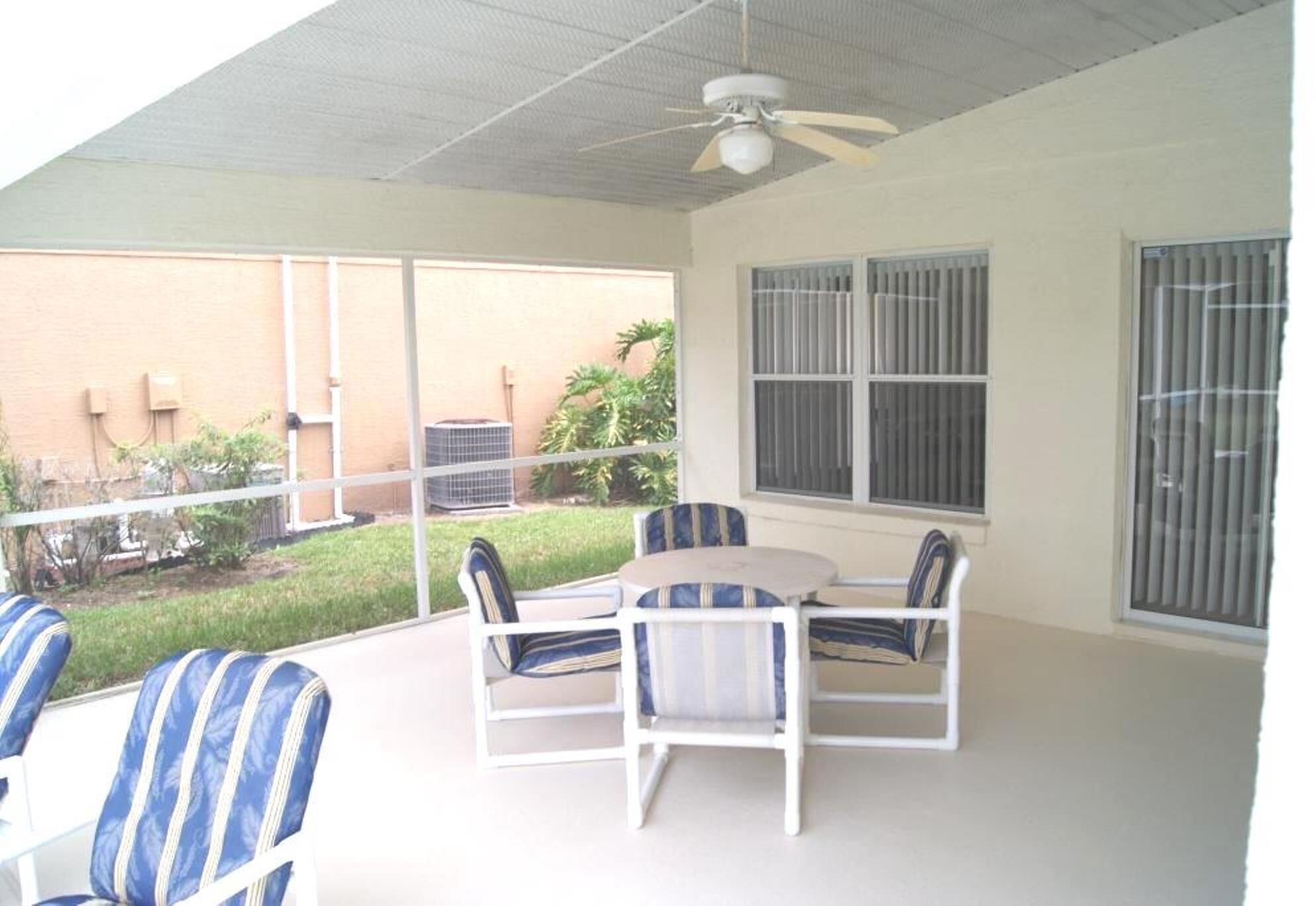 A covered patio with a ceiling fan, white furniture, and large windows offering views of a garden and building exterior.