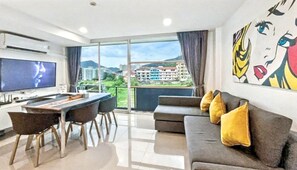 A modern living room with a flat-screen TV, a dining area, a sofa with cushions, and a large window with a city view.