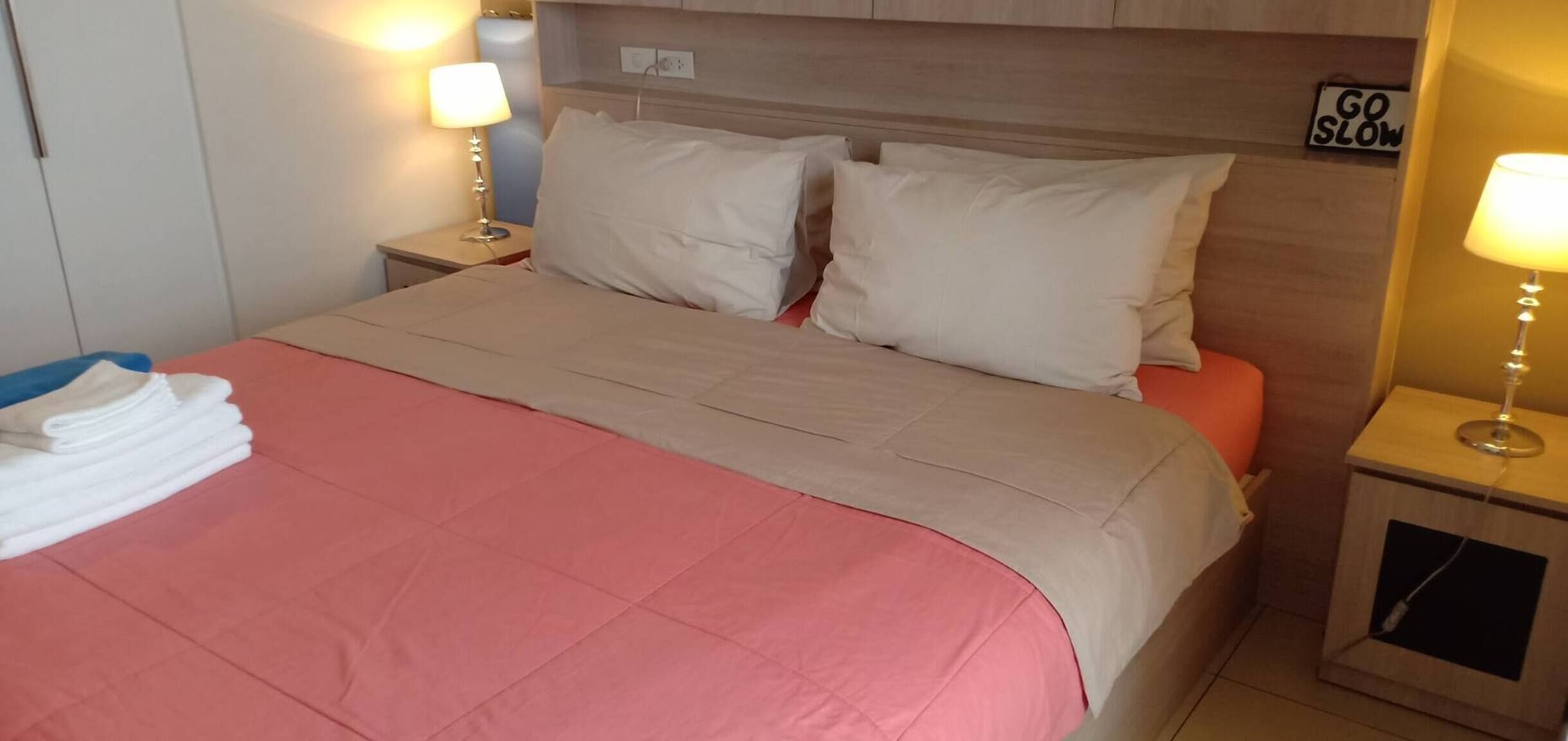 En pænt redt seng med et rosa og beige sengetæppe, to hvide puder, og et skilt på sengegærdet med teksten "GO SLOW".
