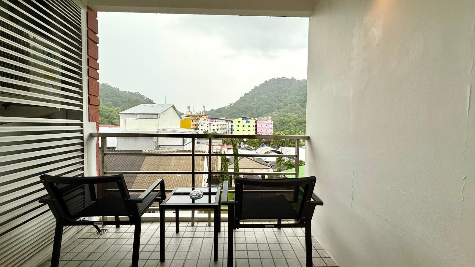 Ein Balkon mit zwei schwarzen Stühlen und einem Tisch, mit Blick auf ein Wohngebiet mit bunten Gebäuden und einen Hügel.