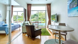 A modern hotel room with a bed, a sofa, a dining table, and a view of a pool and greenery.