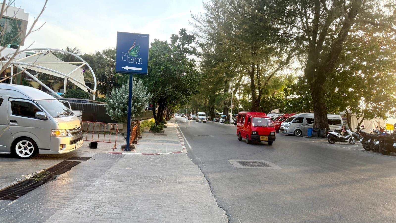 Une vue de rue avec une enseigne indiquant « The Charm Resort Phuket », un trottoir pavé, des voitures garées et des arbres.