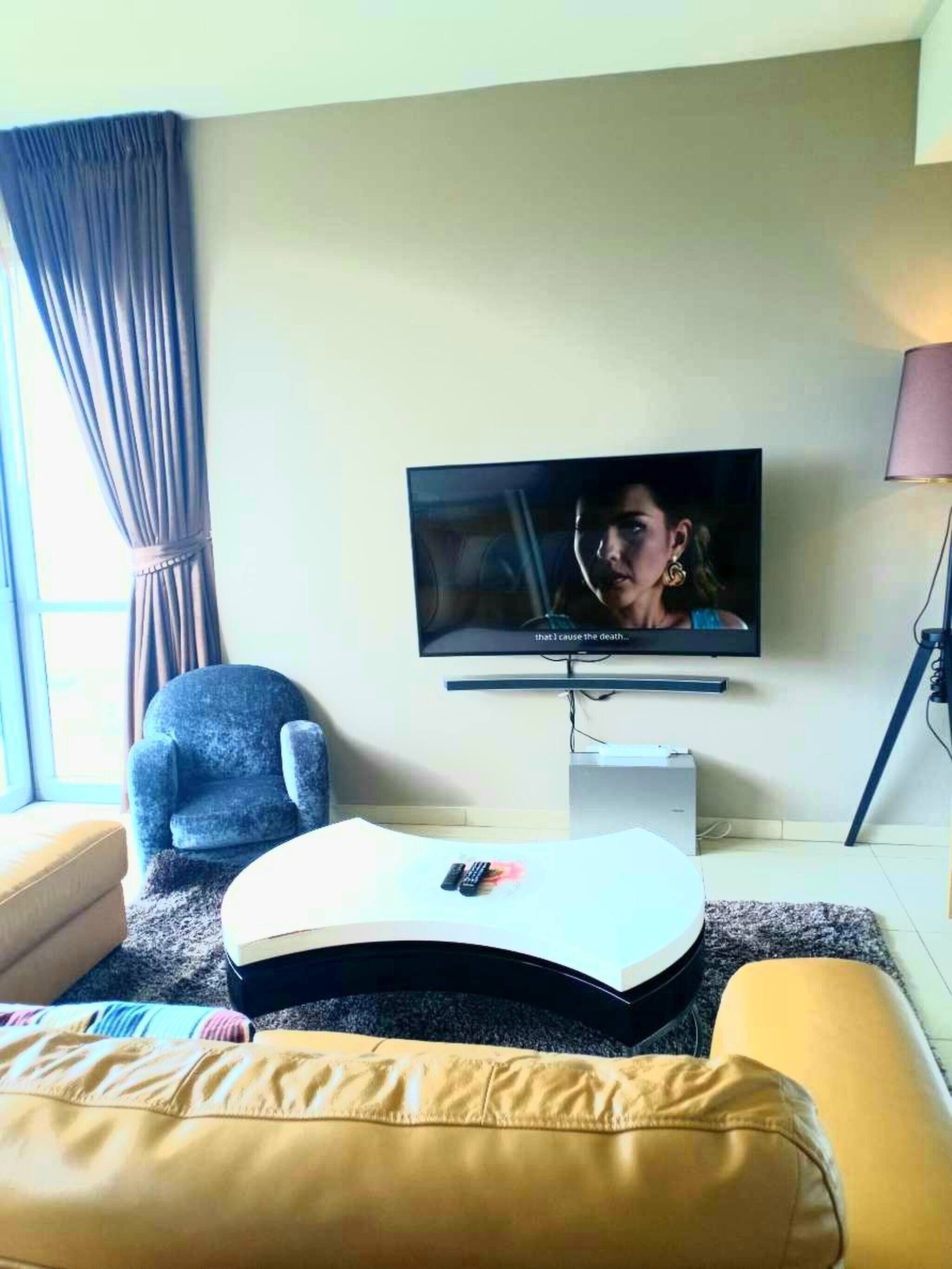 A living room with a flat-screen TV mounted on the wall, a blue armchair, a yellow sofa, and a small white side table with a remote control on it.