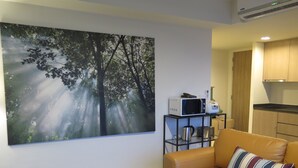 Un salon avec un grand tableau sur le thème de la forêt, un canapé et un petit coin cuisine équipé d’appareils électroménagers.