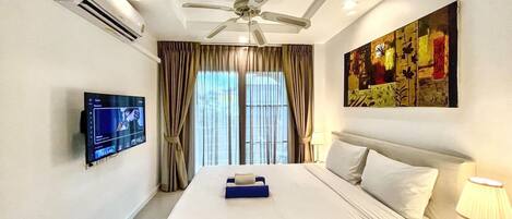 A hotel room with a bed, a ceiling fan, a TV mounted on the wall, and a painting on the wall.