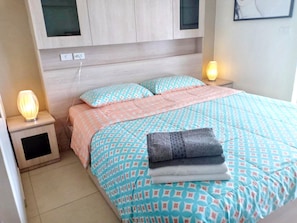 Una cama bien hecha con ropa de cama estampada, una lámpara de noche y una mesita auxiliar pequeña.