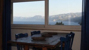 A dining area with a wooden table and chairs, two woven baskets, and a view of a mountainous coastline through large windows. - Casa Gianni Cala Gonone Mare (Sardegna)