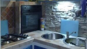 A kitchen with a gas stove, two sinks, a microwave, and a stone wall backsplash. - Casa Gianni Cala Gonone Mare (Sardegna)