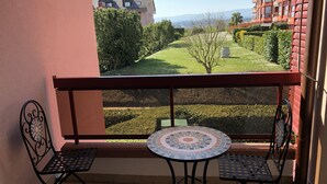 Uma varanda com duas cadeiras de metal e uma mesa pequena, com vista para um jardim bem cuidado e edifícios distantes.