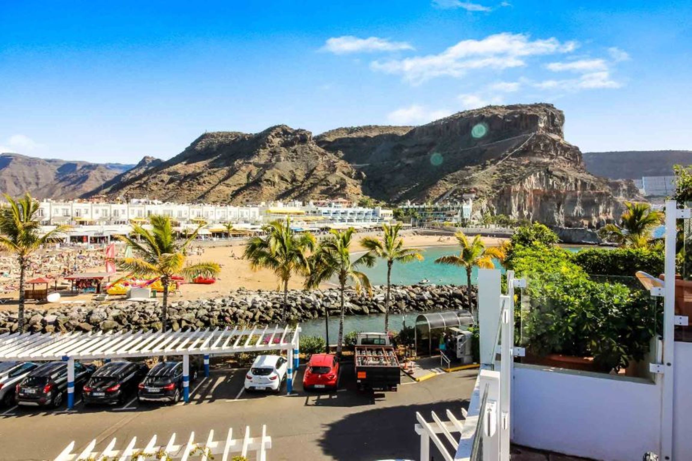 Hotel frente al mar con estacionamiento, playa y montañas al fondo.