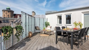 Una terraza en la azotea con una mesa y sillas, rodeada por un recinto de cristal, y una vista de un edificio histórico al fondo.