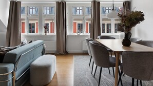 Una sala de estar moderna con un sofá, una mesa de comedor y sillas, ventanas amplias con cortinas y un florero con flores en el alféizar.