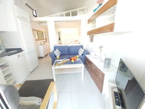 Un espace de vie compact comprenant un coin cuisine, un canapé, une table à manger avec des fruits et des verres, et un téléviseur.