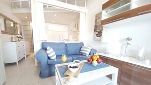 Un salon moderne avec un canapé bleu, une table basse sur laquelle on trouve des fruits et un verre de jus d’orange, et un coin cuisine doté de meubles blancs et d’un plan de travail.
