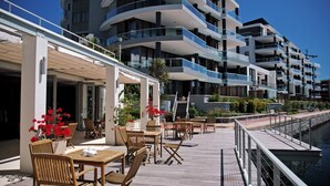 Een terras aan het water met houten tafels en stoelen, een overdekte doorgang en een modern gebouw met balkons op de achtergrond.