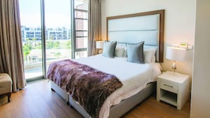 A hotel room with a large bed, a nightstand, a chair, and a view of buildings through the window. - Two Bedroom Apartment - fully furnished and equipped (Cape Town)